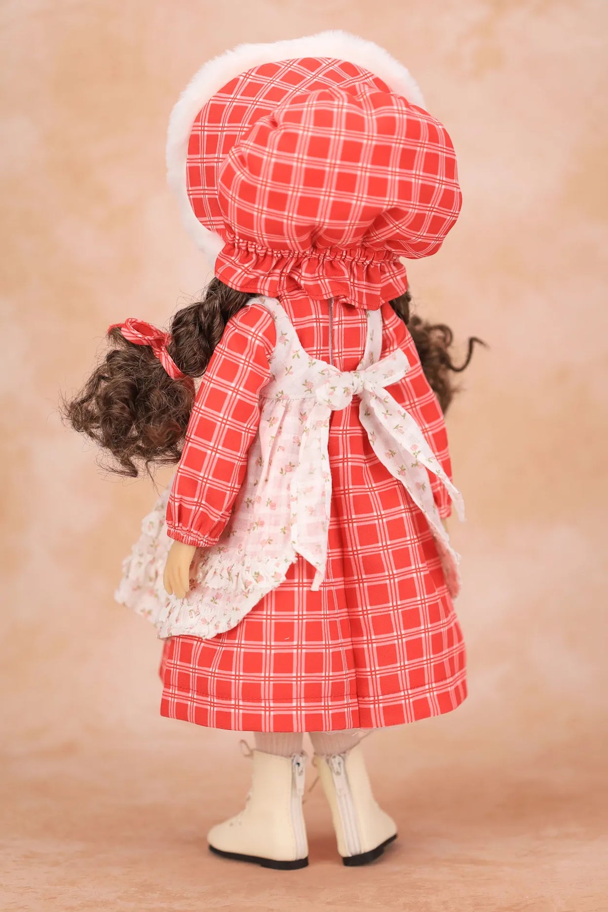 A Holly Hobbie doll viewed from the back wearing a red and white gingham dress with a white lace apron, matching bonnet, and white boots, set against a warm beige background.