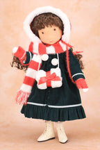 A Holly Hobbie doll dressed in a dark green dress with a red and white striped scarf, a matching red and white striped bonnet with white trim, and cream-colored boots, set against a soft beige background.
