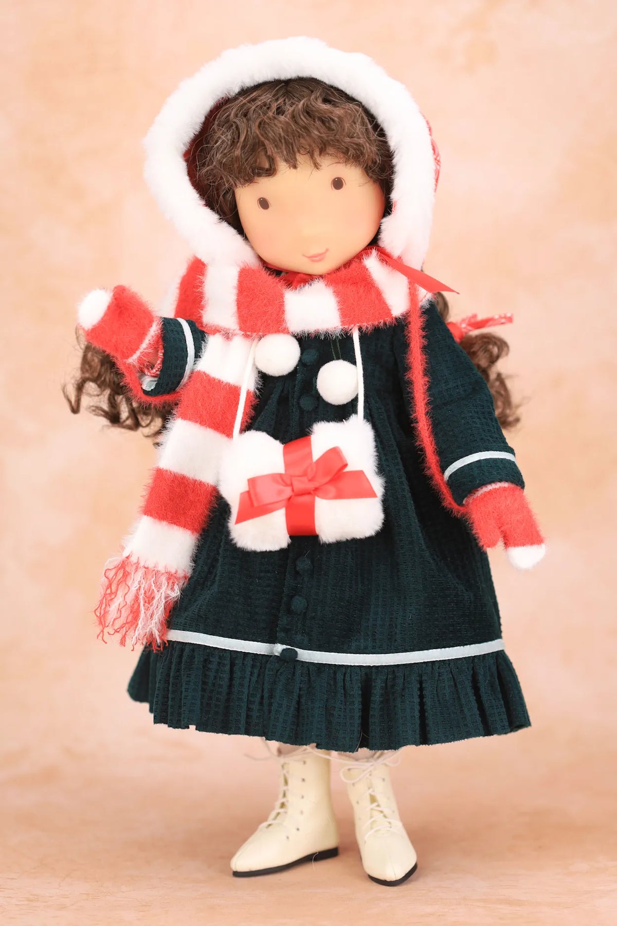 A Holly Hobbie doll dressed in a dark green dress with a red and white striped scarf, a matching red and white striped bonnet with white trim, and cream-colored boots, set against a soft beige background.