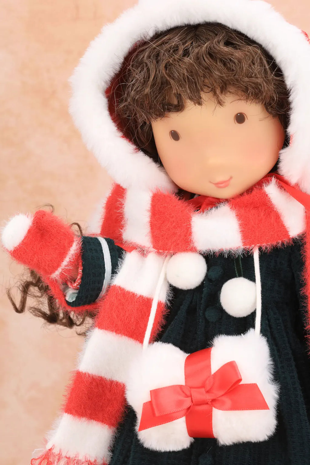 A close-up of a Holly Hobbie doll wearing a festive winter outfit with a red and white striped hooded scarf, a dark green dress against a soft beige background.