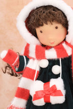A close-up of a Holly Hobbie doll wearing a festive winter outfit with a red and white striped hooded scarf, a dark green dress against a soft beige background.