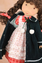 A close-up of a Holly Hobbie doll with brown curly hair wearing a dark green jacket with white pom-poms, a pink floral dress, and a red plaid skirt against a soft beige background.