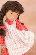 A close-up of a Holly Hobbie doll with long brown curly hair wearing a red plaid shirt and a white floral pinafore dress, posed against a soft beige background.