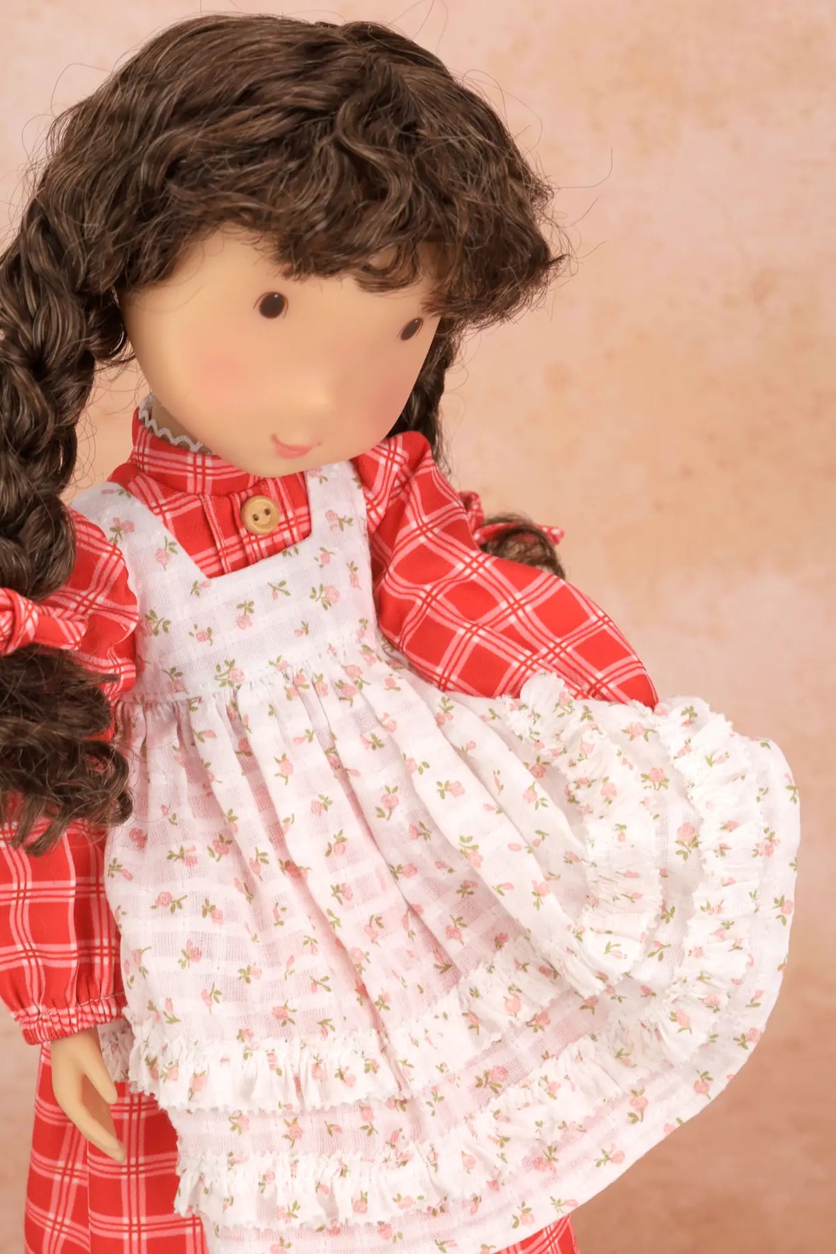 A close-up of a Holly Hobbie doll with long brown curly hair wearing a red plaid shirt and a white floral pinafore dress, posed against a soft beige background.