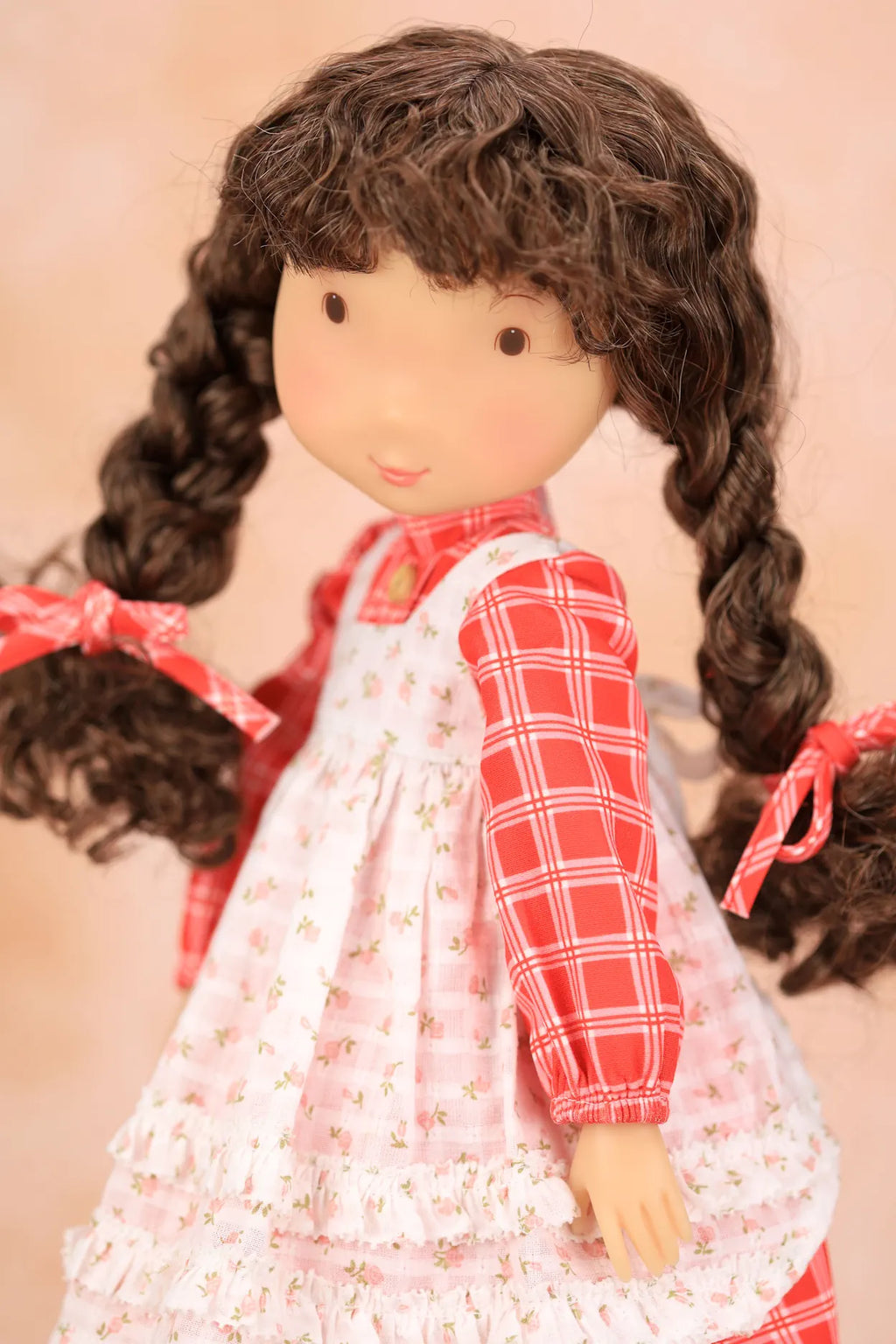 A close-up of a Holly Hobbiedoll with long dark brown curly hair in pigtails, wearing a red and white plaid shirt under a white floral pinafore dress, with pink bows in her hair, set against a soft peach background.