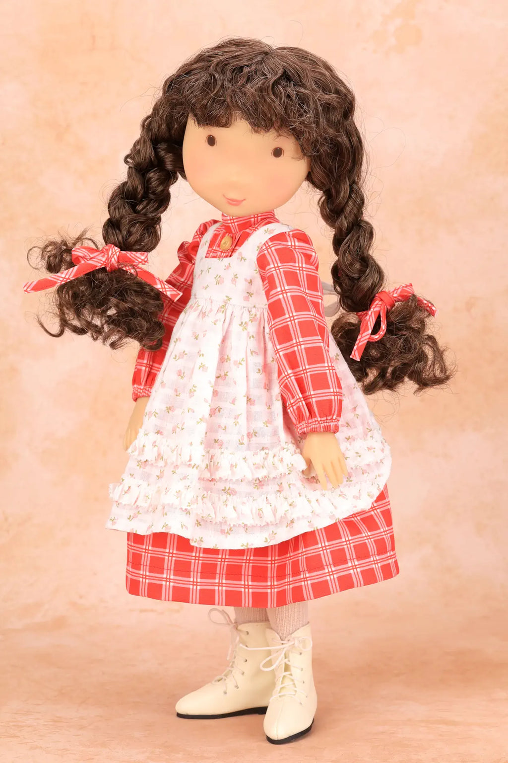 A Holly Hobbie doll with long dark curly hair styled in two braids, wearing a red and white plaid dress over a white floral apron, with cream-colored boots, set against a warm beige background.