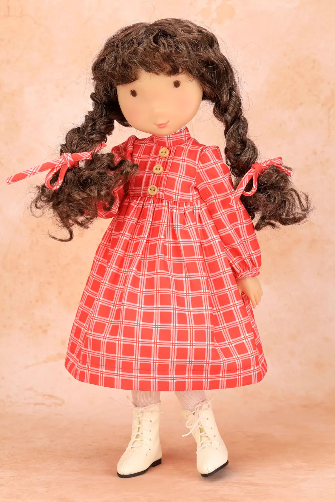 A vintage-style Holly Hobbie doll with long dark curly hair tied in pigtails, wearing a red and white plaid dress with cream lace-up boots, posed against a warm beige textured background.