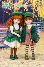 Two dolls with red hair and green outfits standing in a scenic outdoor setting.