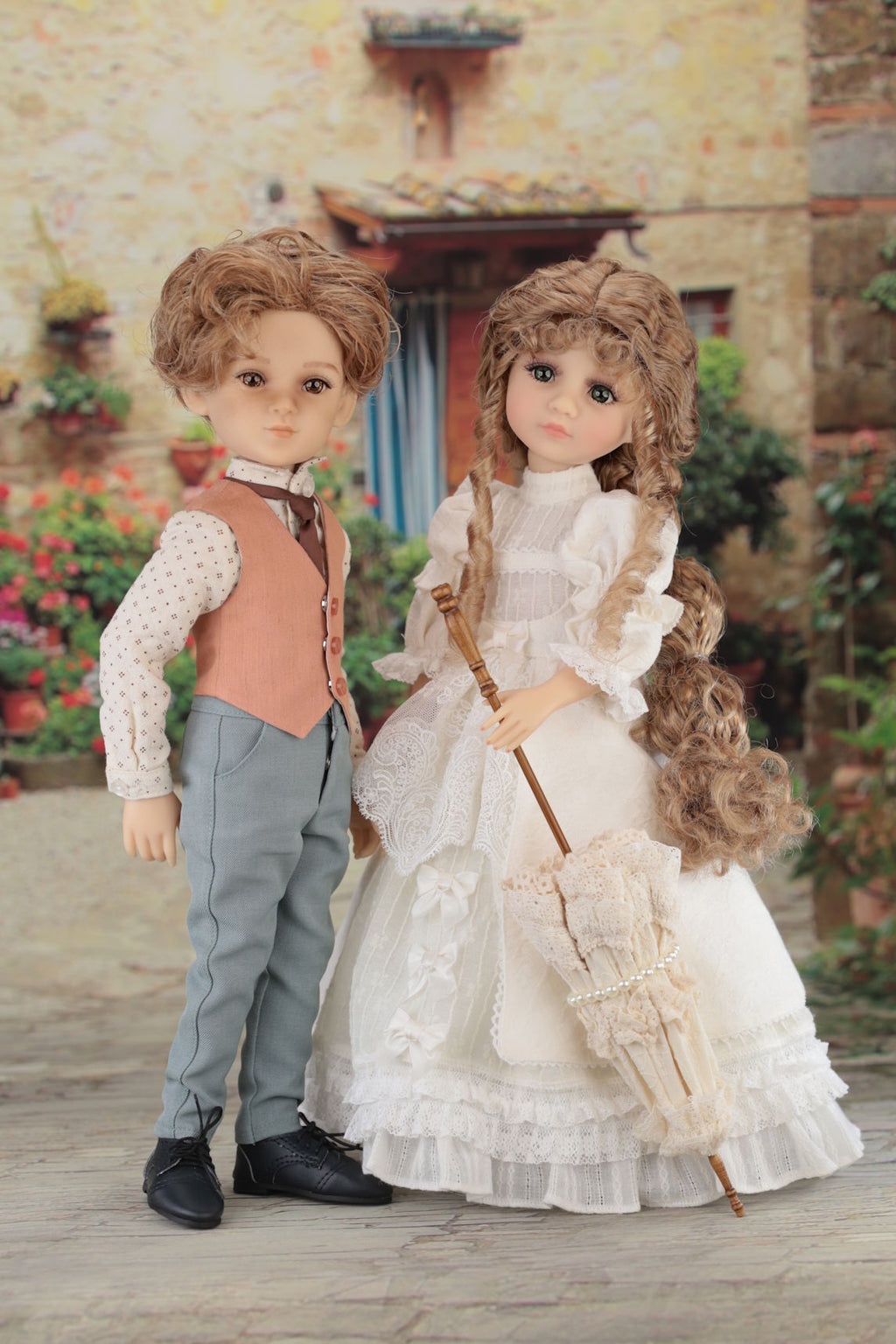 Two dolls, a boy and a girl, standing together in an outdoor setting with flowers and a building in the background.