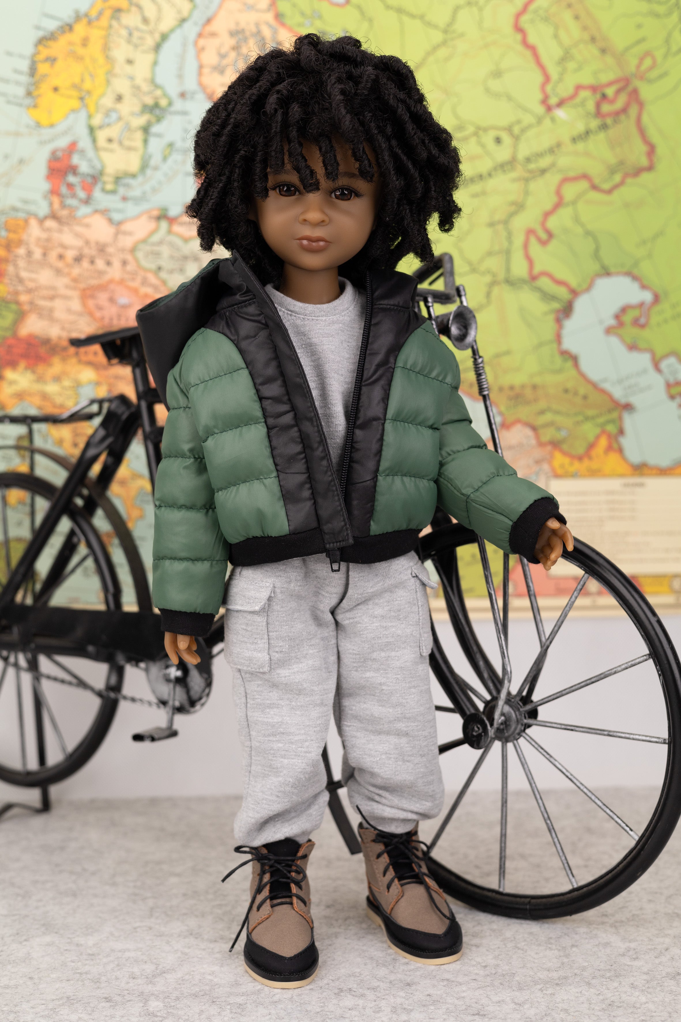 Doll in green puffer jacket and gray pants standing next to a bicycle with a world map in the background