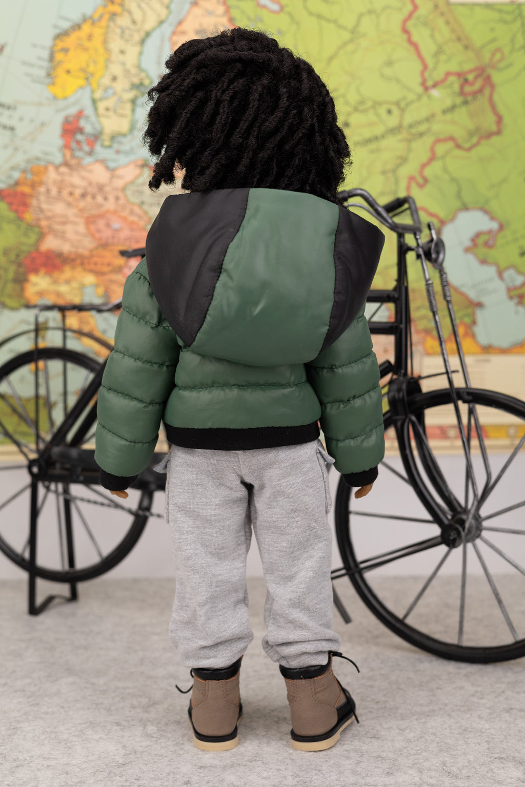 Doll wearing a green and black puffer jacket standing in front of a bicycle and world map.