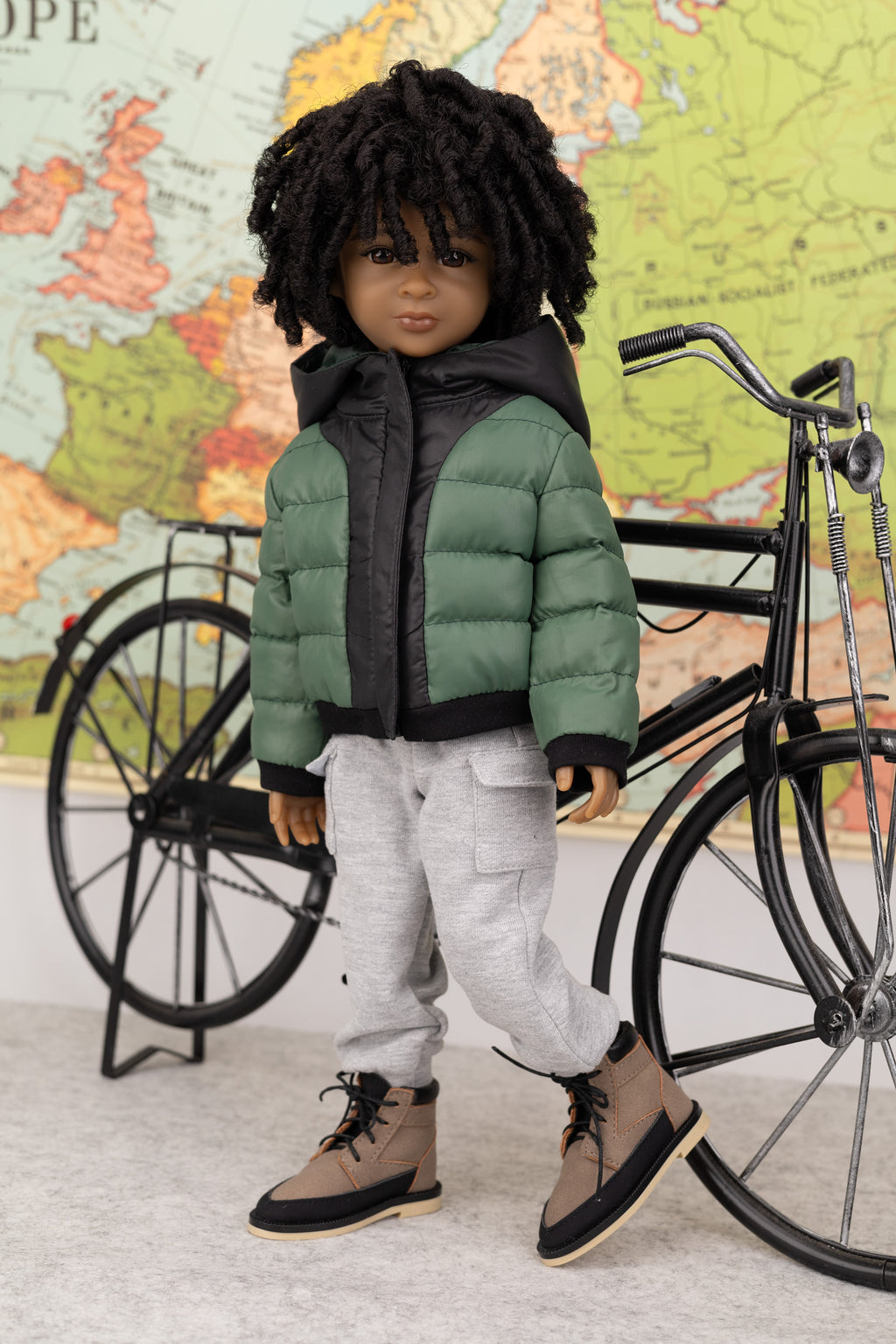 Doll wearing a green puffer jacket and gray pants, standing next to a bicycle with a world map in the background.