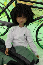 Doll wearing a gray sweatshirt with a small black logo, sitting on a green cushion with a bicycle in the background.