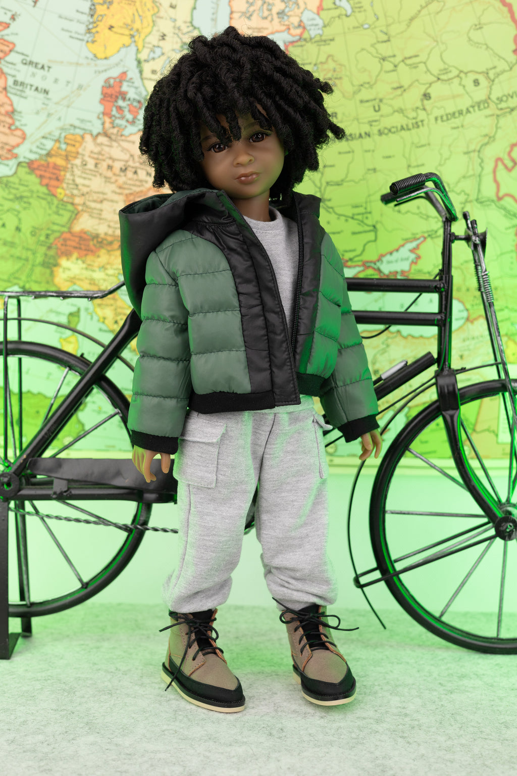 Doll wearing a green puffer jacket and gray pants standing next to a bicycle with a world map in the background