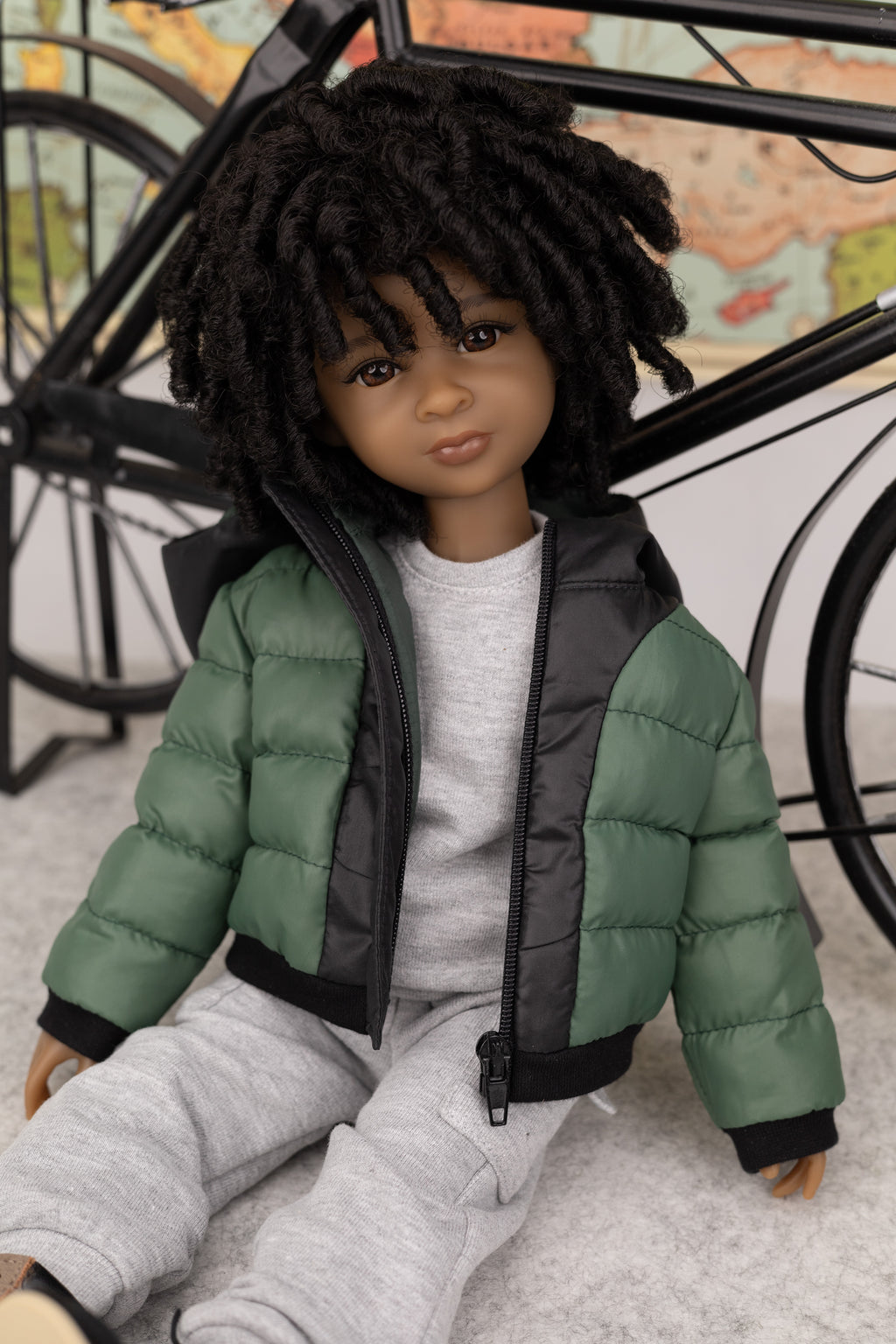 Doll wearing a green puffer jacket and gray pants sitting on a surface with a blurred background.