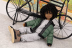 Doll wearing a green jacket and gray pants, sitting next to a bicycle with a world map in the background.