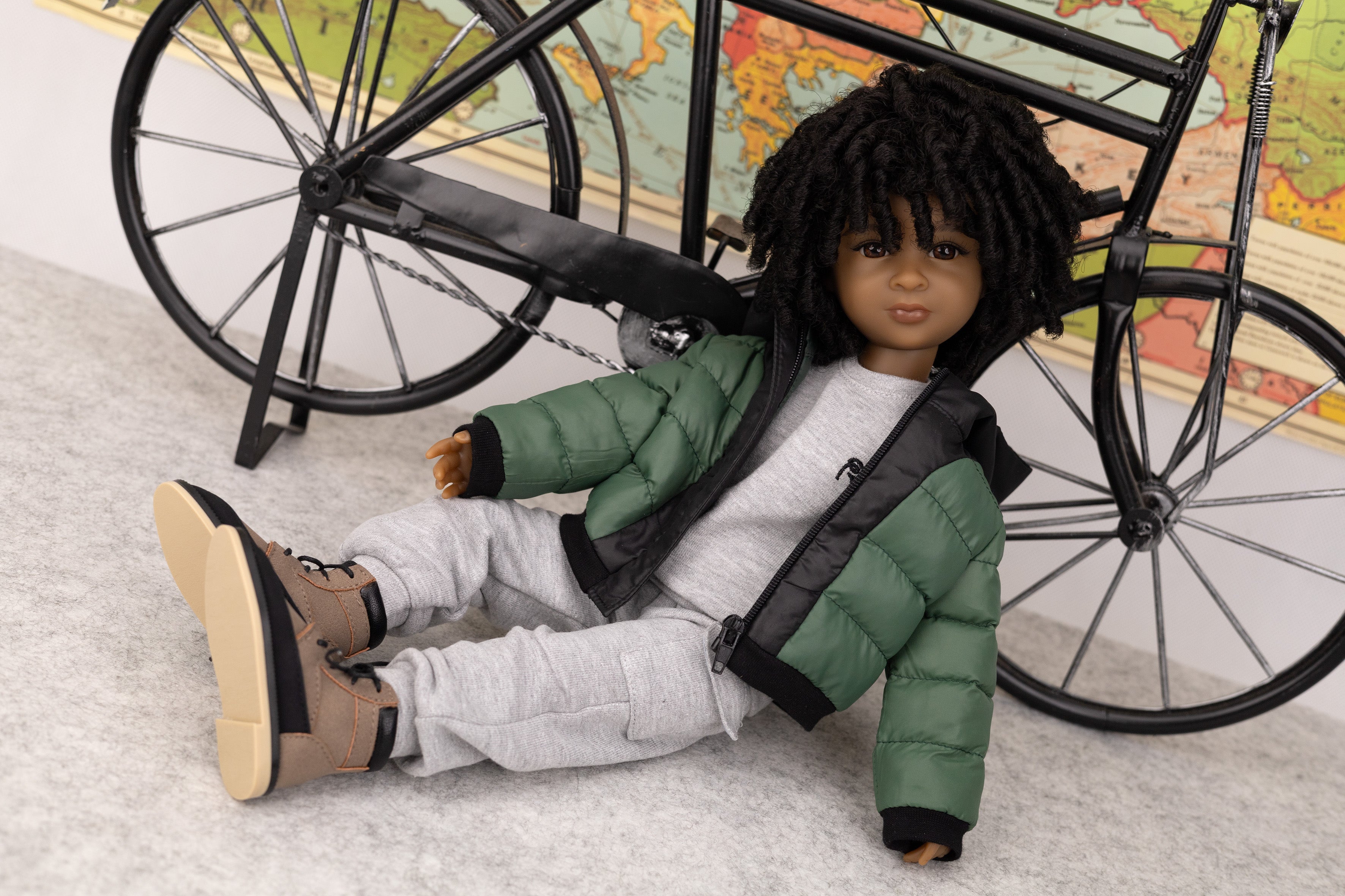 Doll wearing a green jacket and gray pants, sitting next to a bicycle with a world map in the background.