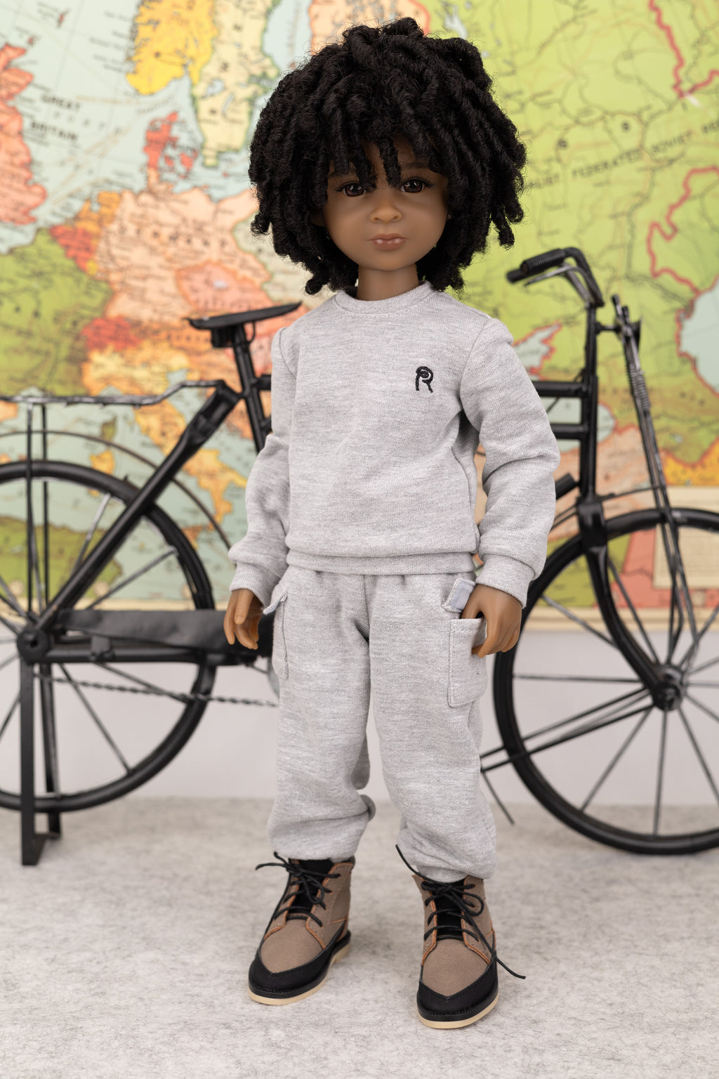 Doll in gray outfit standing next to a bicycle with a world map in the background