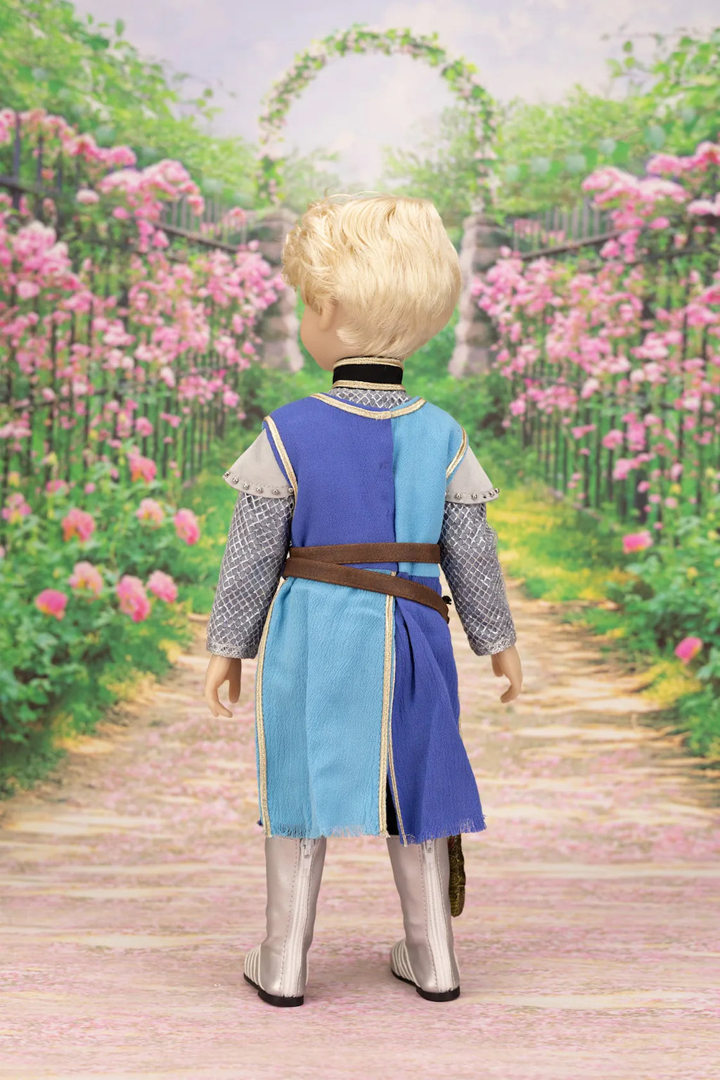 Lancelot doll in a costume standing on a path with pink flowers and an archway in the background