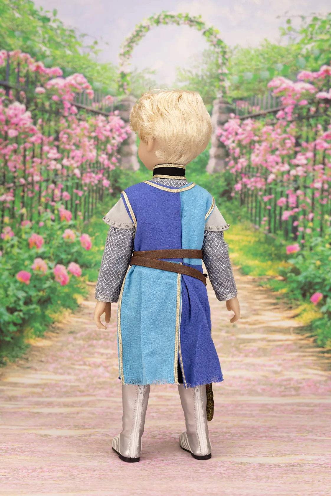 Lancelot doll in a costume standing on a path with pink flowers and an archway in the background