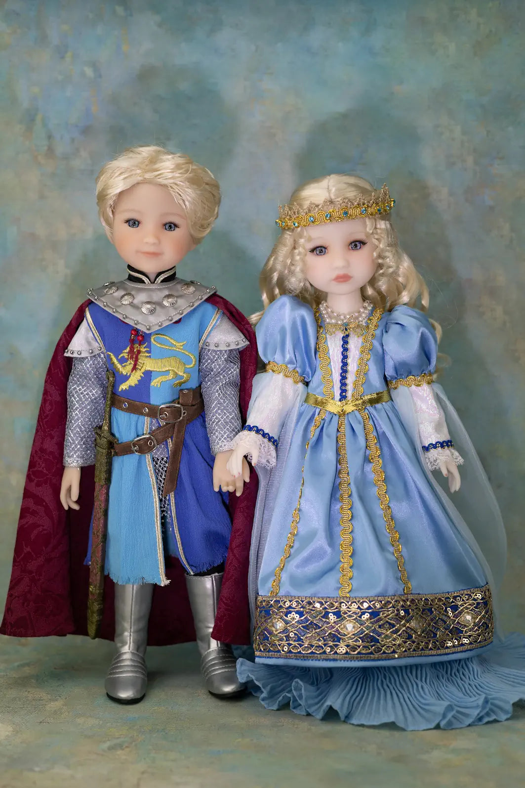 Two dolls dressed in medieval royal costumes standing side by side against a textured blue studio backdrop. The doll on the left wears a blonde wig, silver armor, a blue tunic, brown belt, and red cape.