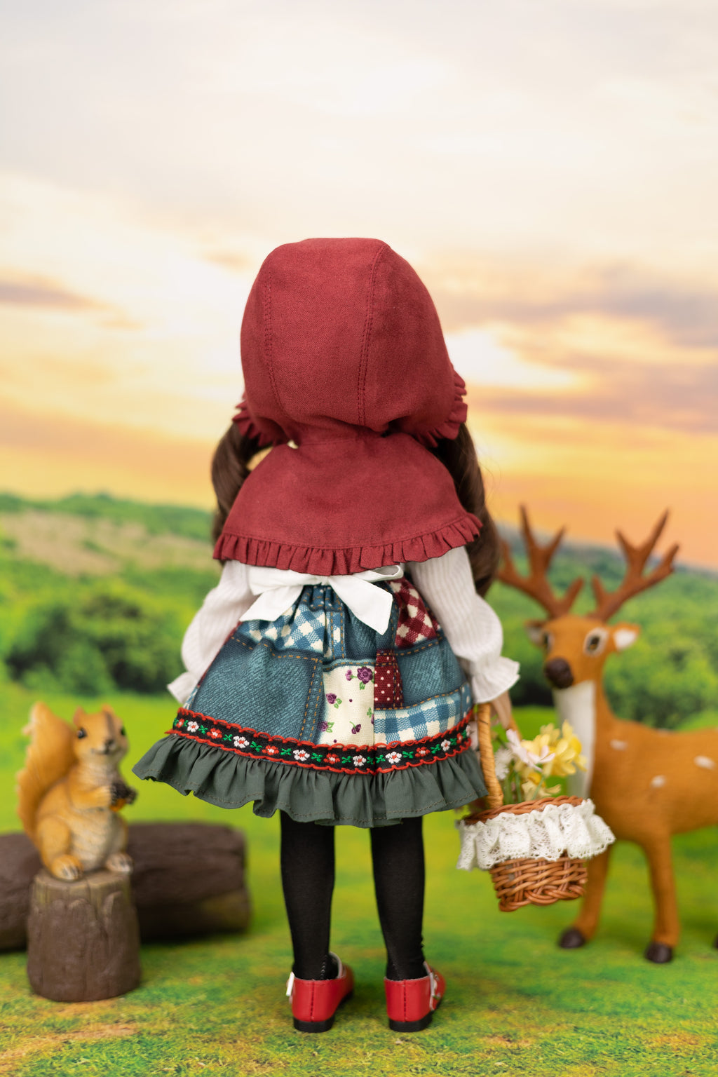 Doll in a red hood and checkered dress standing in a scenic outdoor setting with a deer and squirrel.