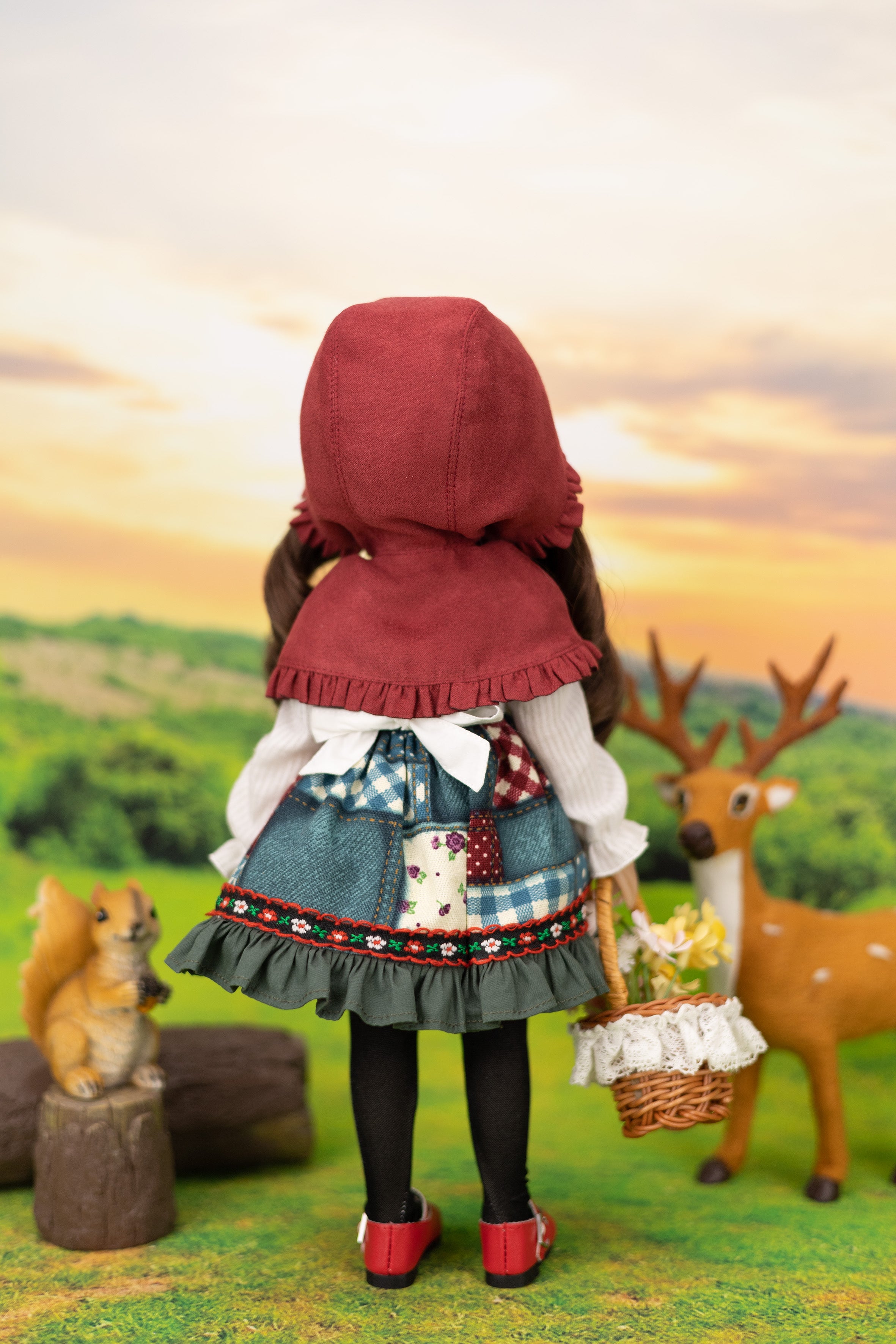 Doll in a red hood and checkered dress standing in a scenic outdoor setting with a deer and squirrel.