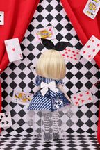 Doll with blonde hair and a blue dress standing on a checkered floor with playing cards around.