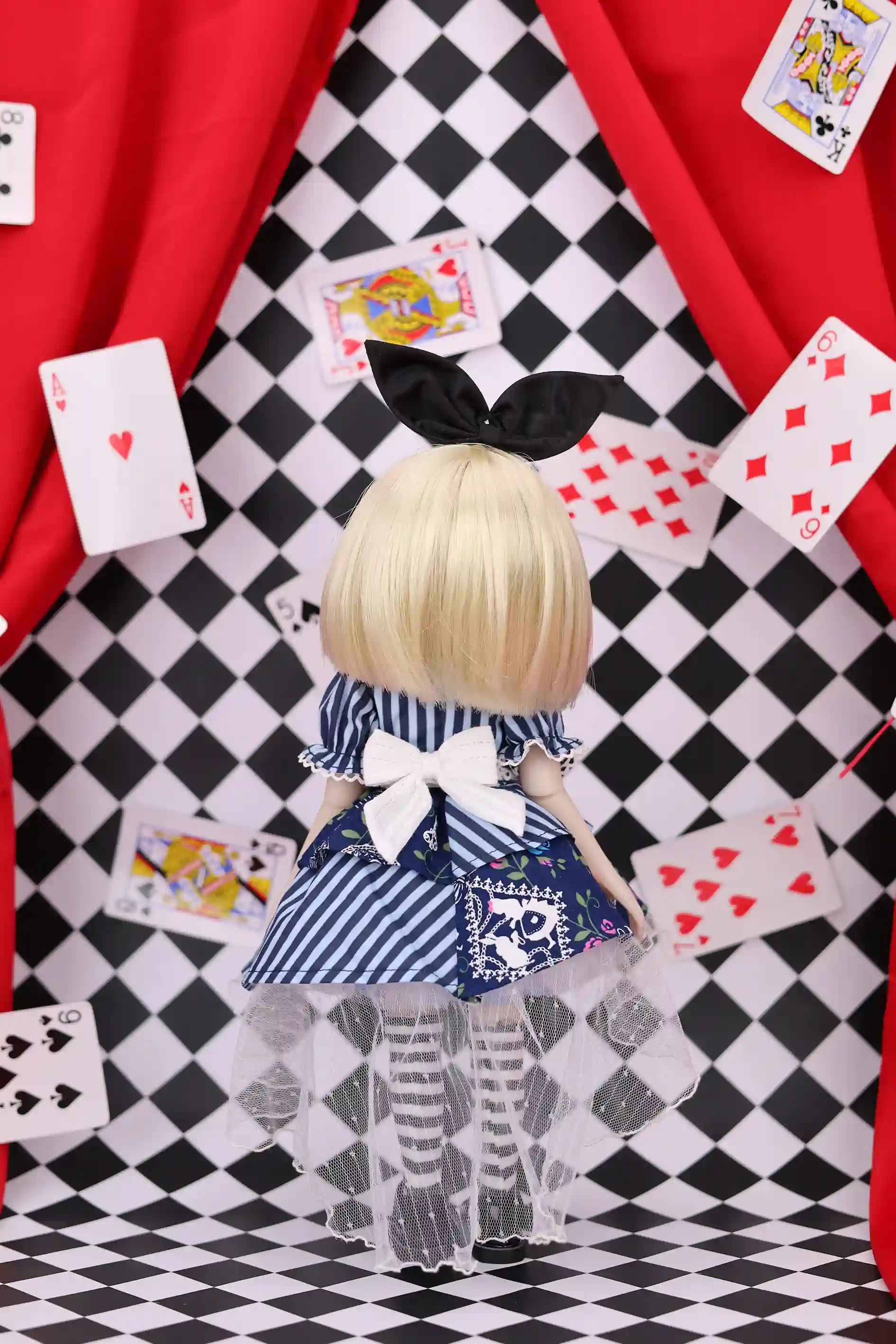 Doll with blonde hair and a blue dress standing on a checkered floor with playing cards around.