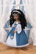 Doll in a blue and white dress with jewelry, standing on a wooden floor with white curtains in the background.