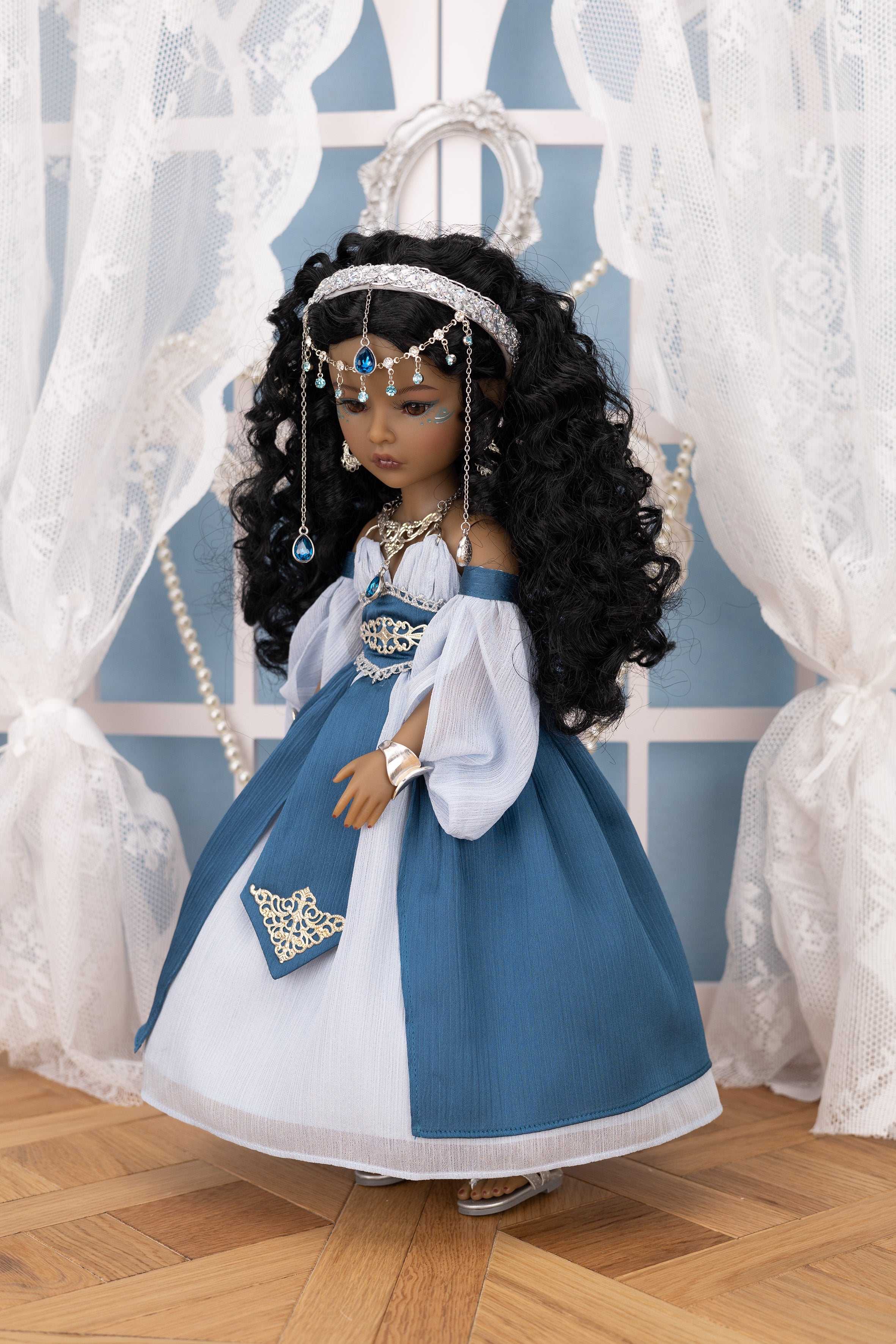 Doll in a blue and white dress with jewelry, standing on a wooden floor with white curtains in the background.
