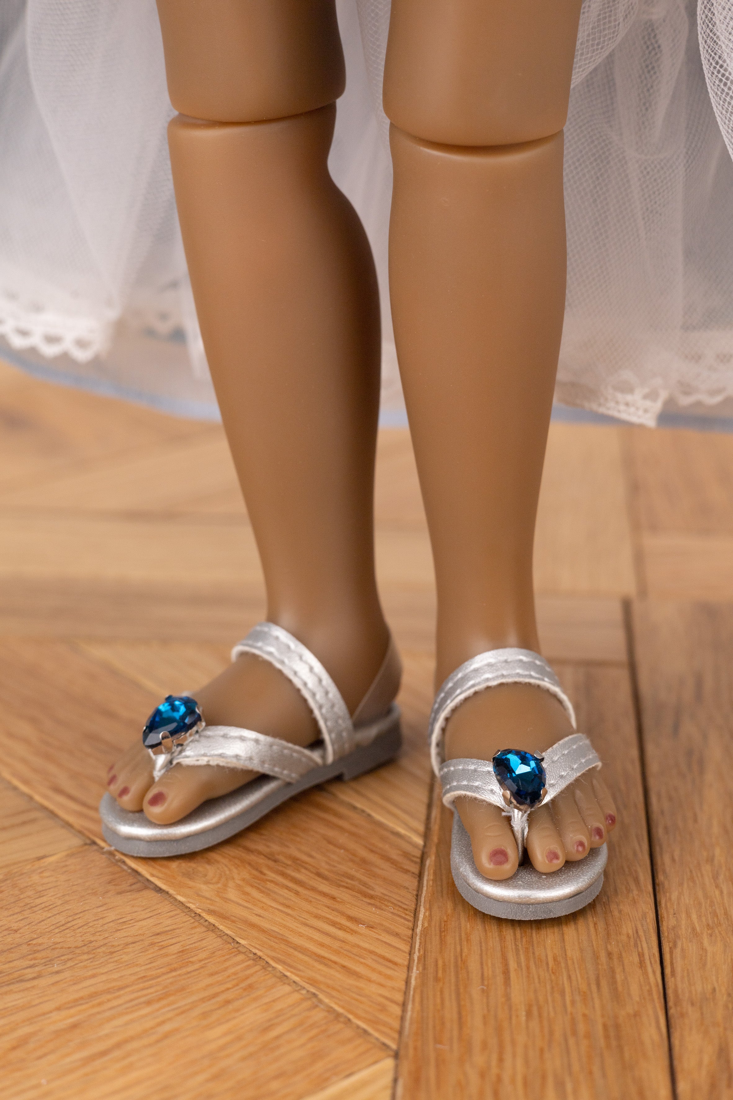 Close-up of doll feet wearing silver sandals with blue stones on a wooden floor.