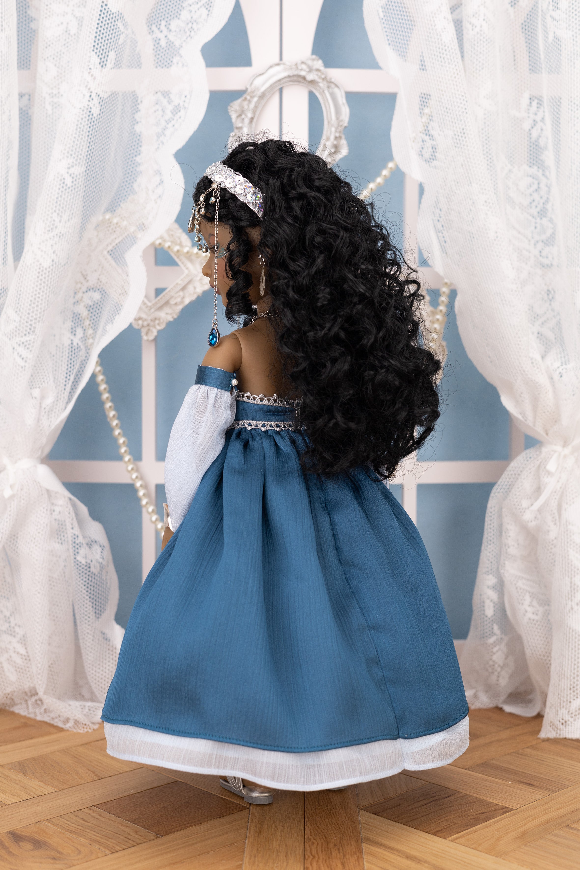 Doll in a blue dress with a tiara against a decorative white curtain background