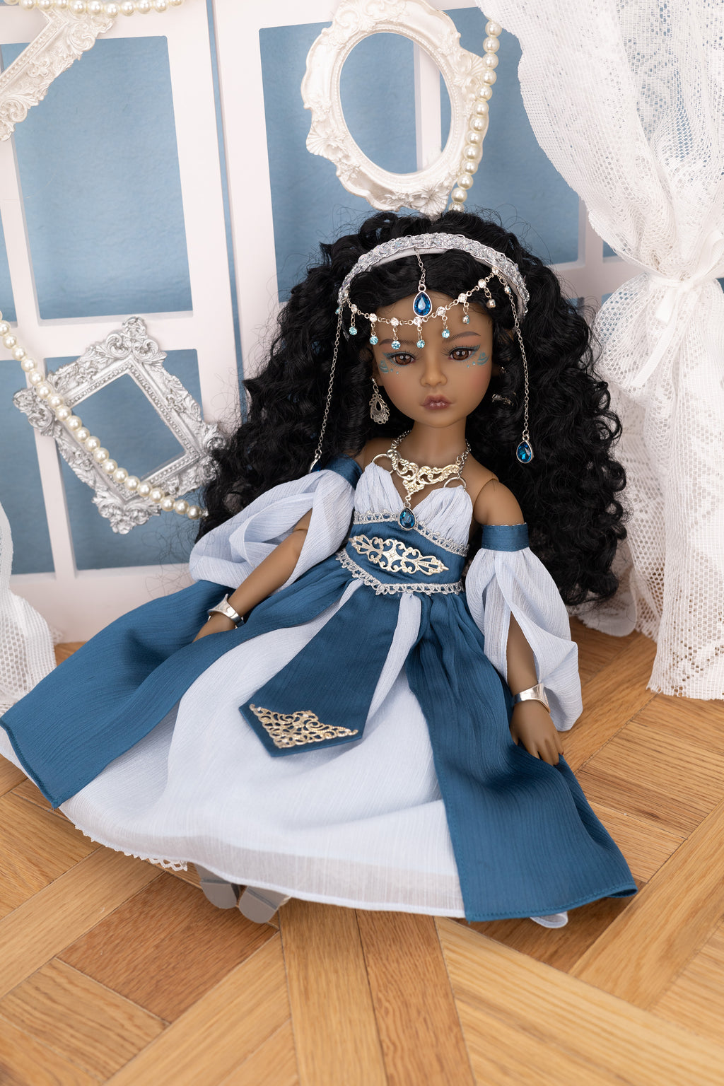 Doll in a blue and white dress with jewelry on a wooden floor.