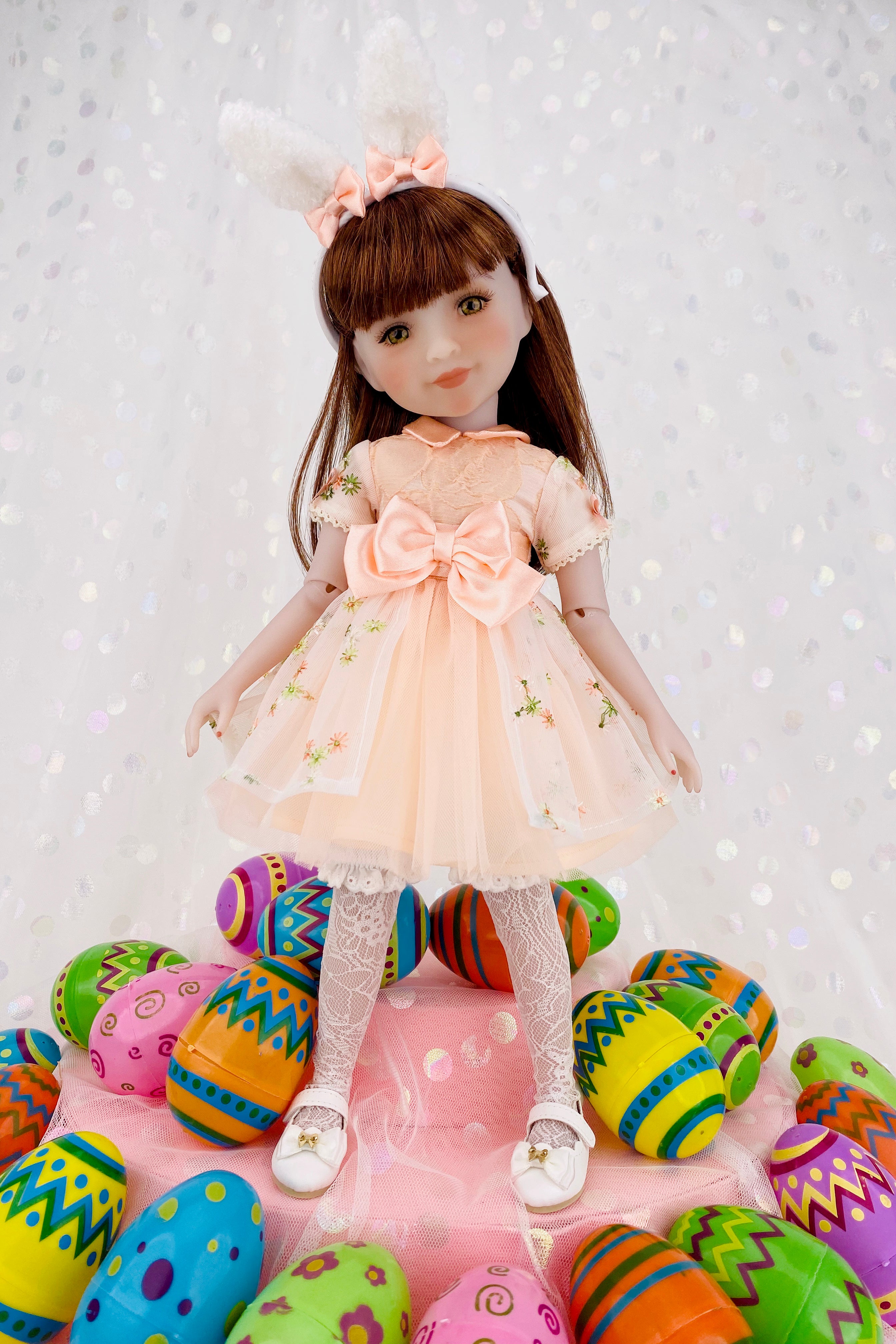 Doll in a light dress with bunny ears surrounded by colorful Easter eggs on a white background