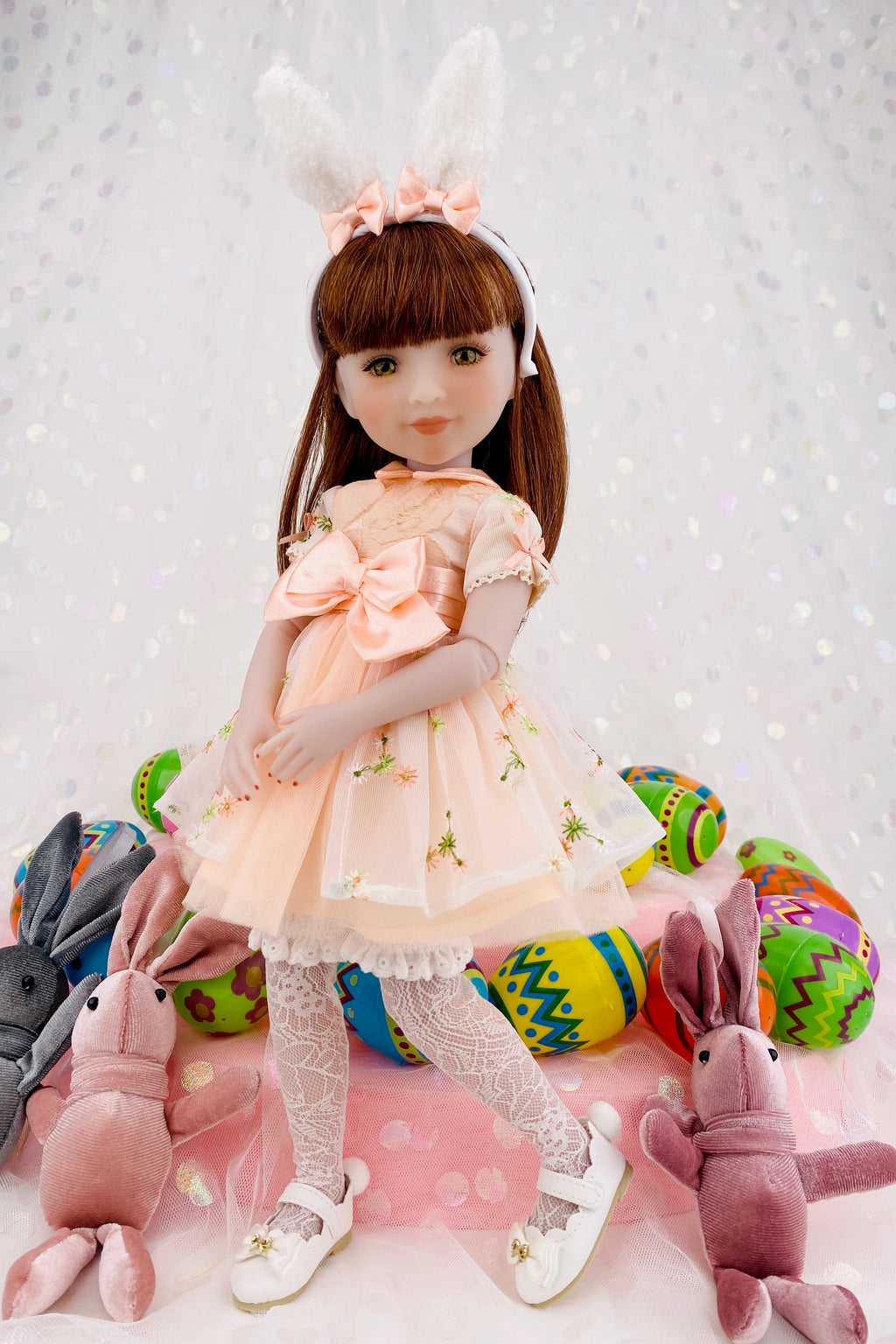 Doll in a light dress with bunny ears surrounded by colorful Easter eggs and plush toys on a soft pink background.