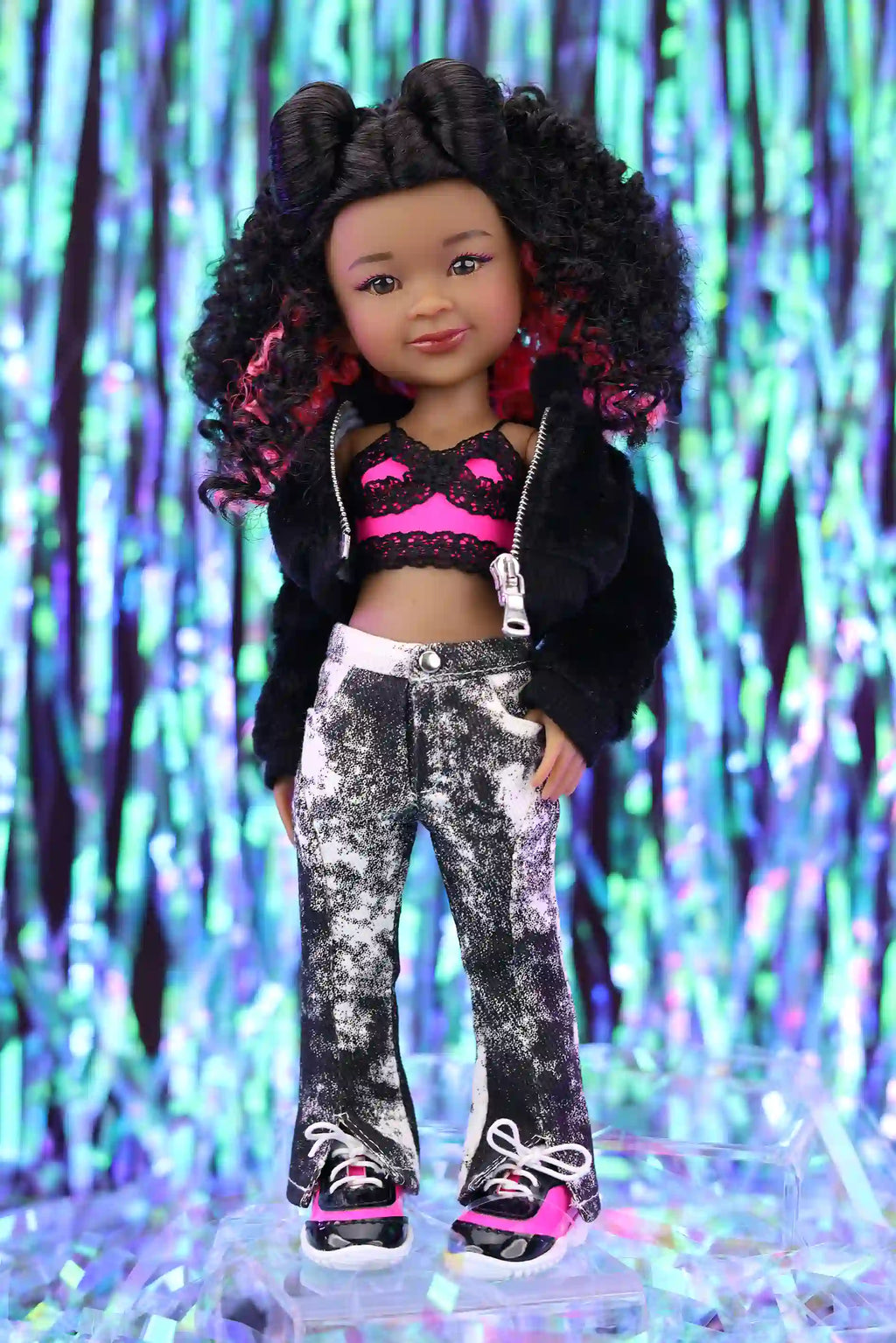 Doll wearing a black jacket, pink top, and patterned pants with a colorful, blurred background.