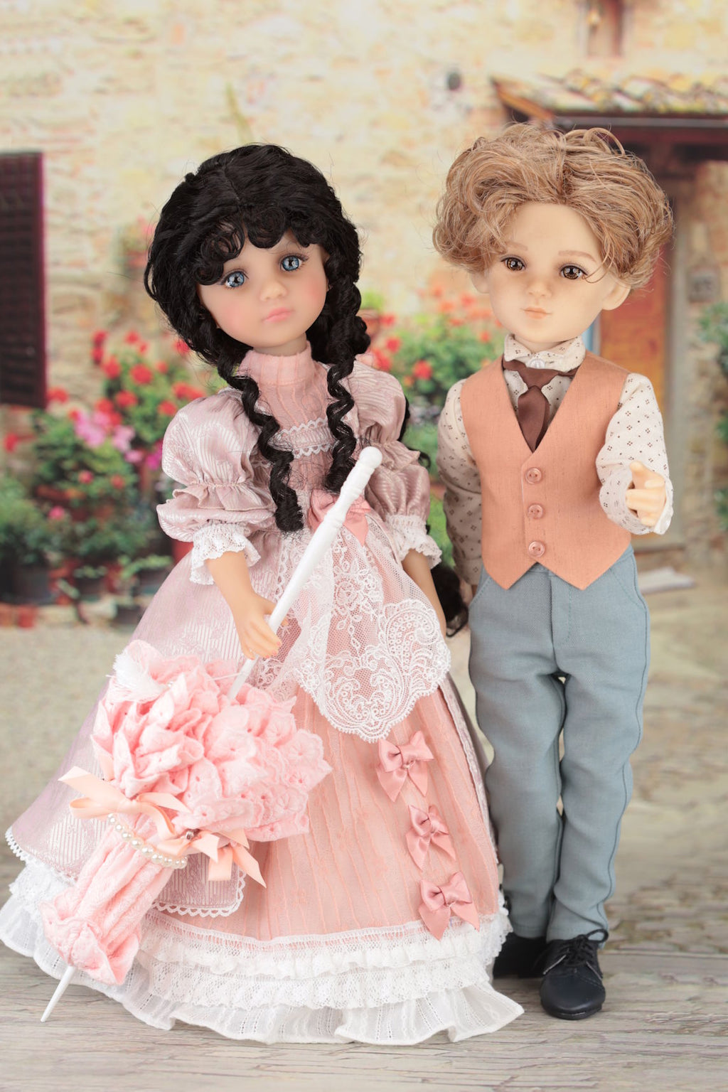 Two dolls, one in a pink dress and the other in formal attire, standing outdoors with flowers in the background.