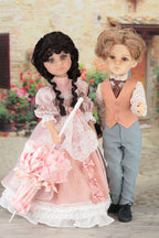 Two dolls, one in a pink dress and the other in formal attire, standing outdoors with flowers in the background.