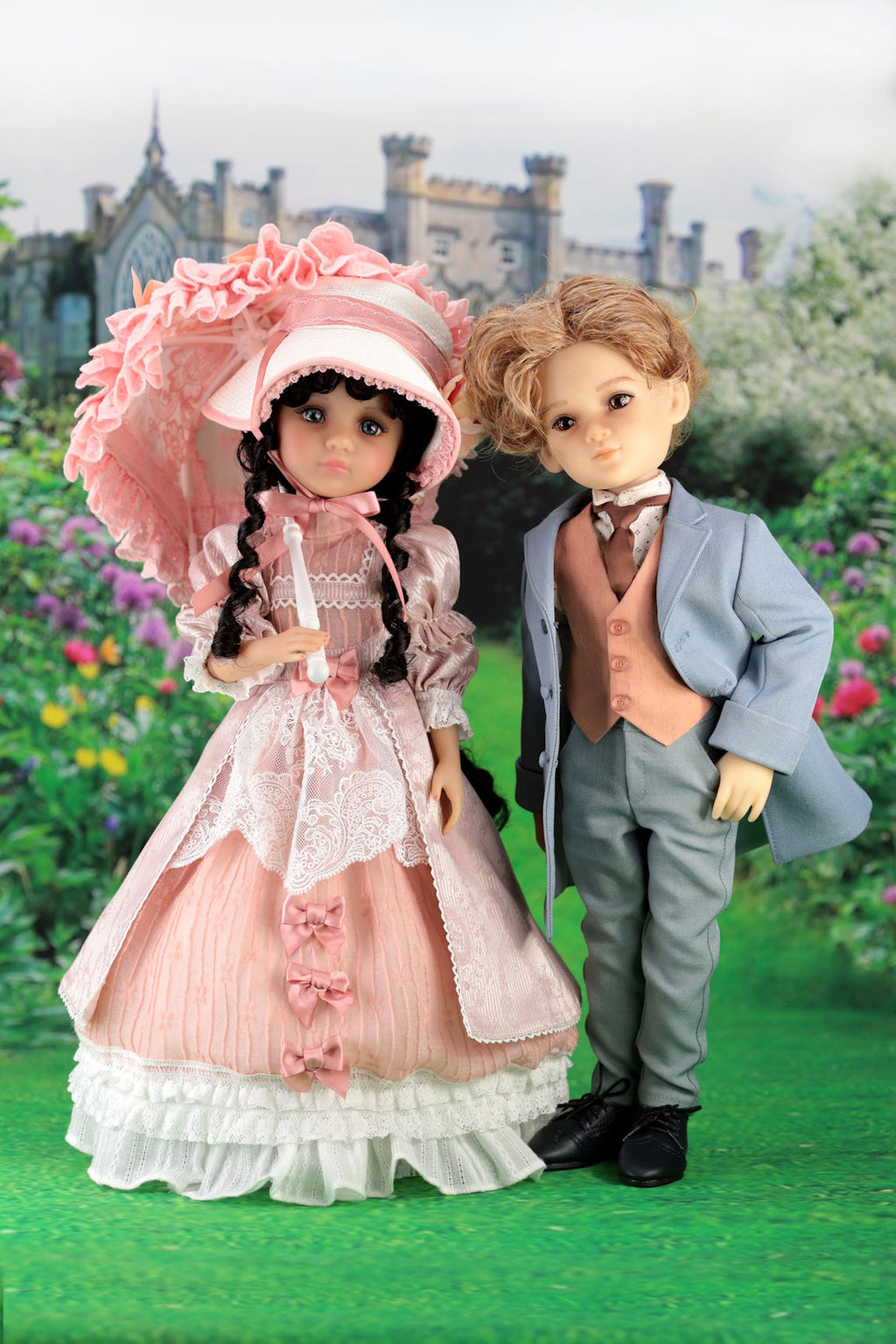 Two dolls, one in a pink dress and hat and the other in a formal outfit, standing in front of a castle.