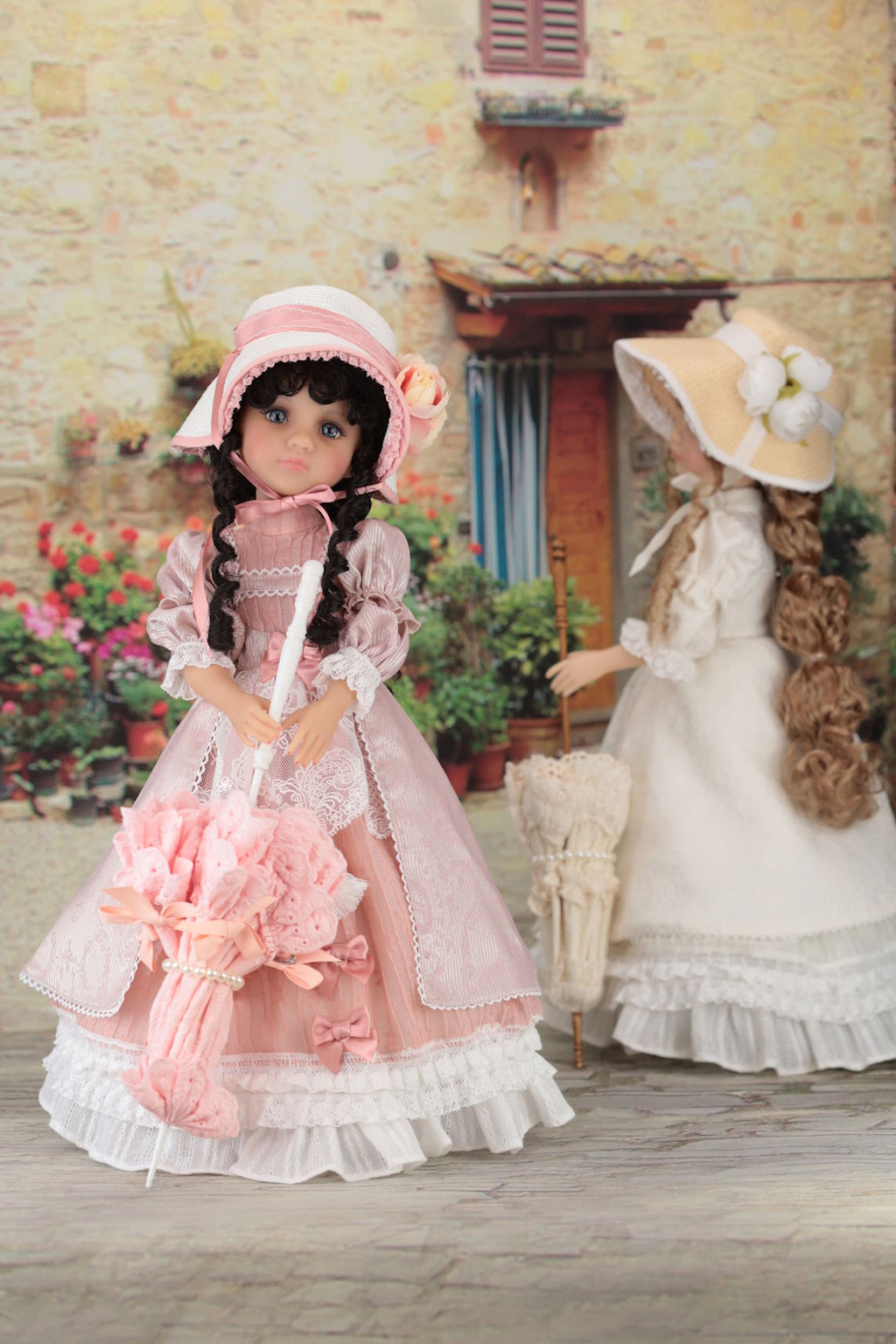 Two dolls in vintage dresses and hats standing in front of a brick building with flowers.