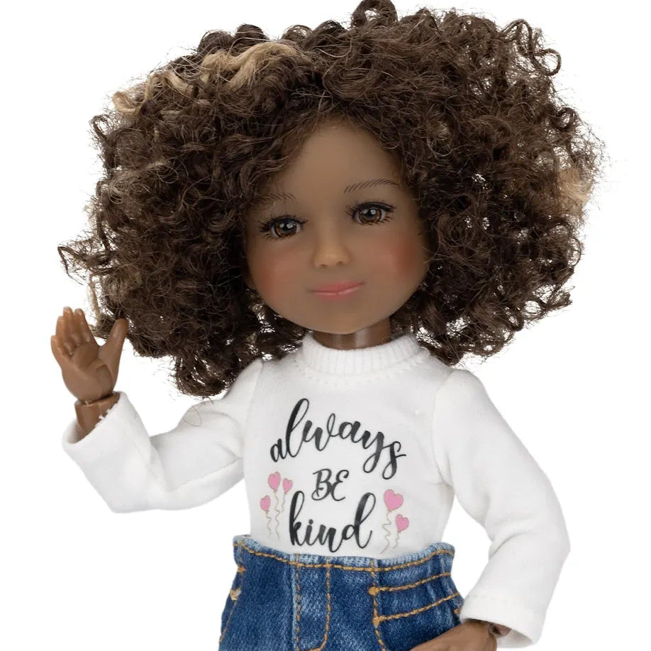 A doll with dark brown curly hair wearing a white long-sleeve shirt with 'always be kind' printed on the front, a blue denim skirt, patterned leggings, and patterned shoes, set against a plain white background.