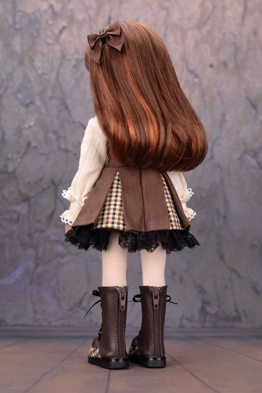 Back view of a doll with long brown hair, wearing a brown outfit with a checkered skirt against a textured gray wall.