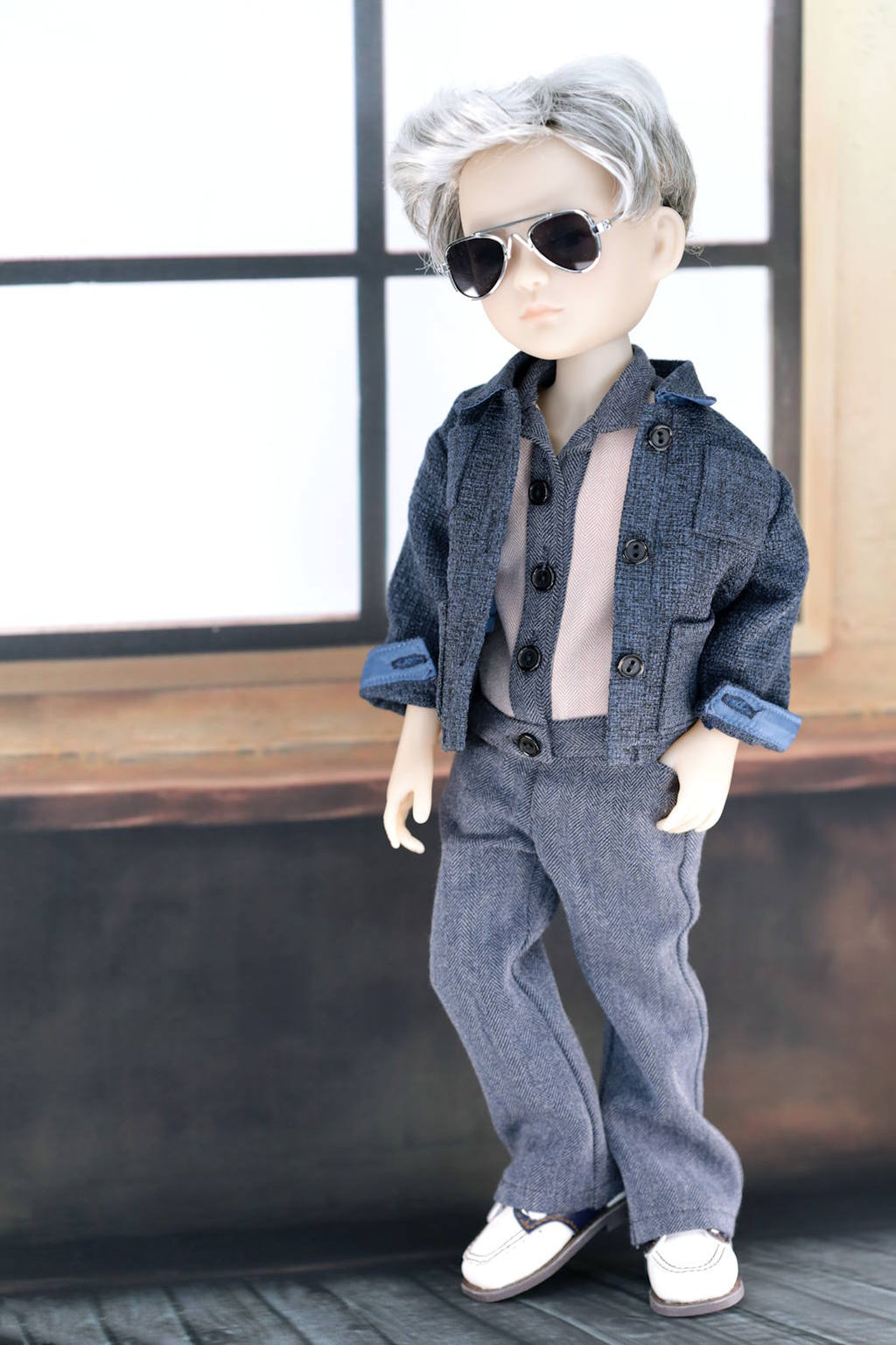 Doll wearing a blue outfit with sunglasses in a room with large windows.