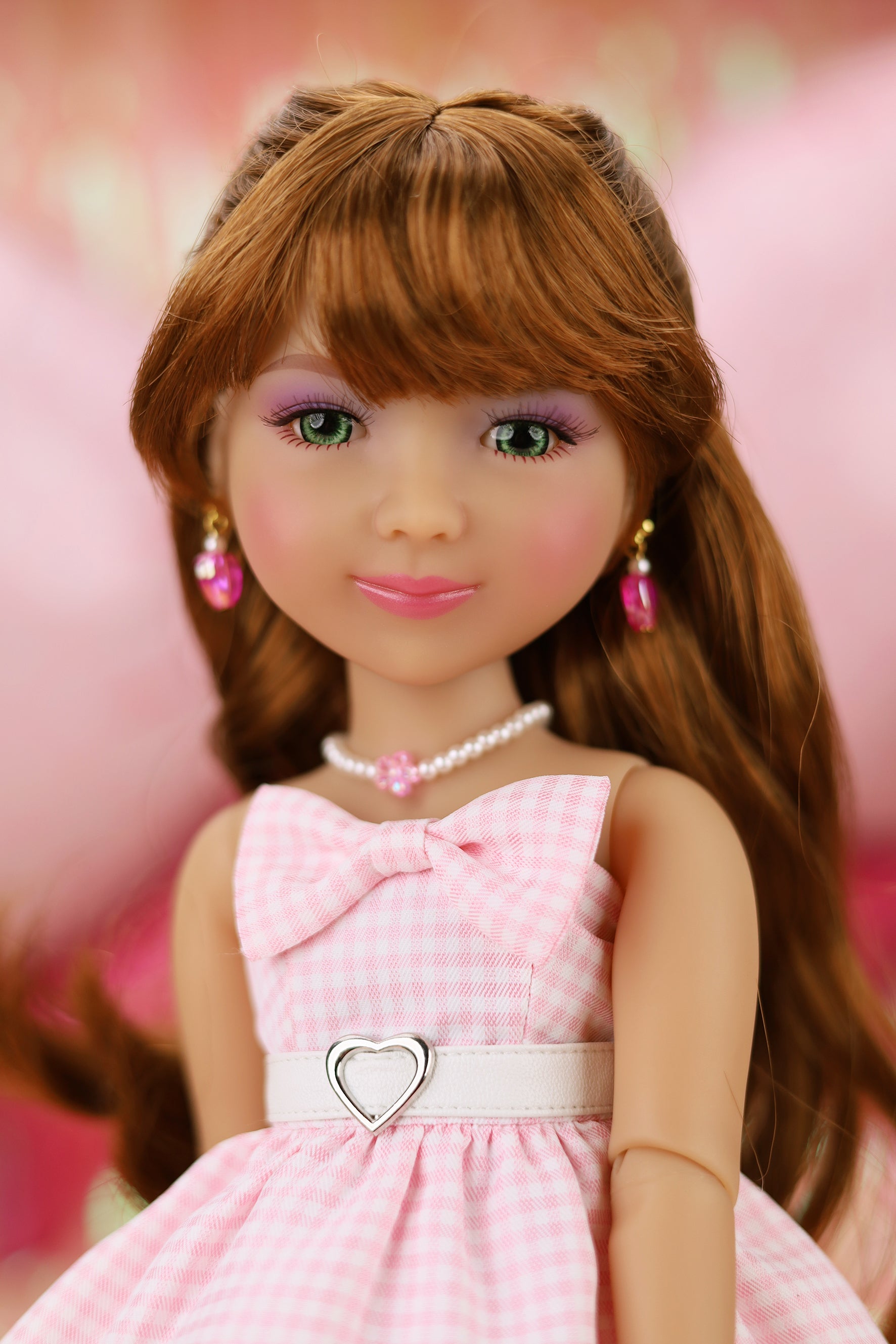 Doll with long brown hair wearing a pink dress with a bow and heart-shaped belt against a pink background.