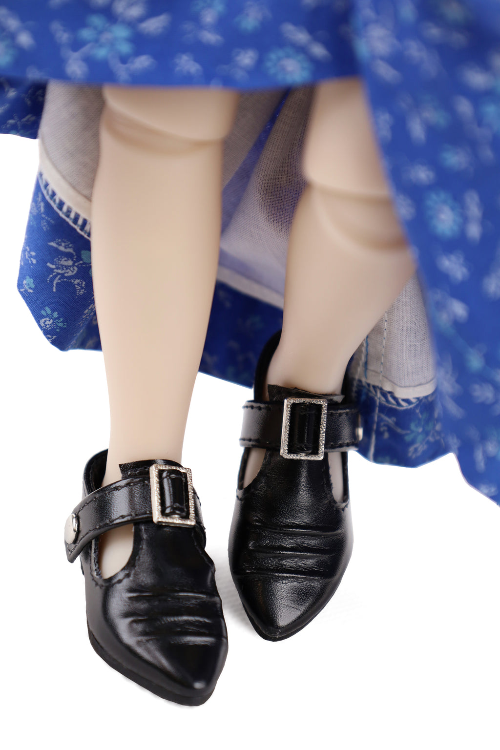 Black doll shoes with buckles worn by a figure in a blue floral dress on a white background