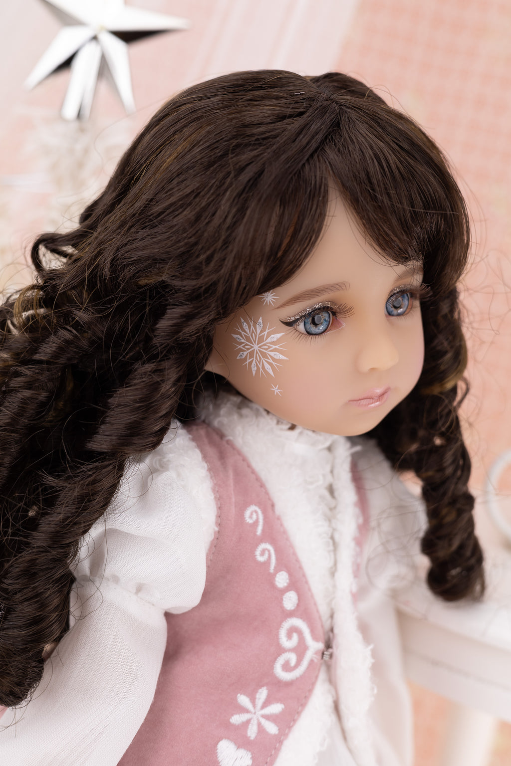 Doll with long brown hair styled in pigtails, wearing a white blouse and pink vest with floral patterns on a soft pink background.