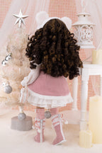 Doll in a pink dress with white fur trim standing in front of a Christmas tree.