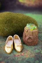 Gold shoes with white pearls and a frog on a wooden log against a green textured background