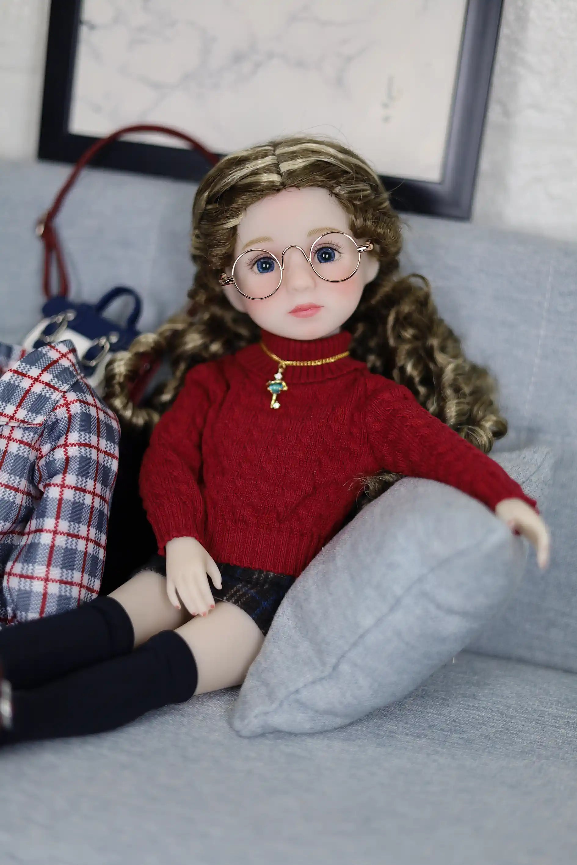 Doll wearing a red sweater and glasses sitting on a couch with a pillow.